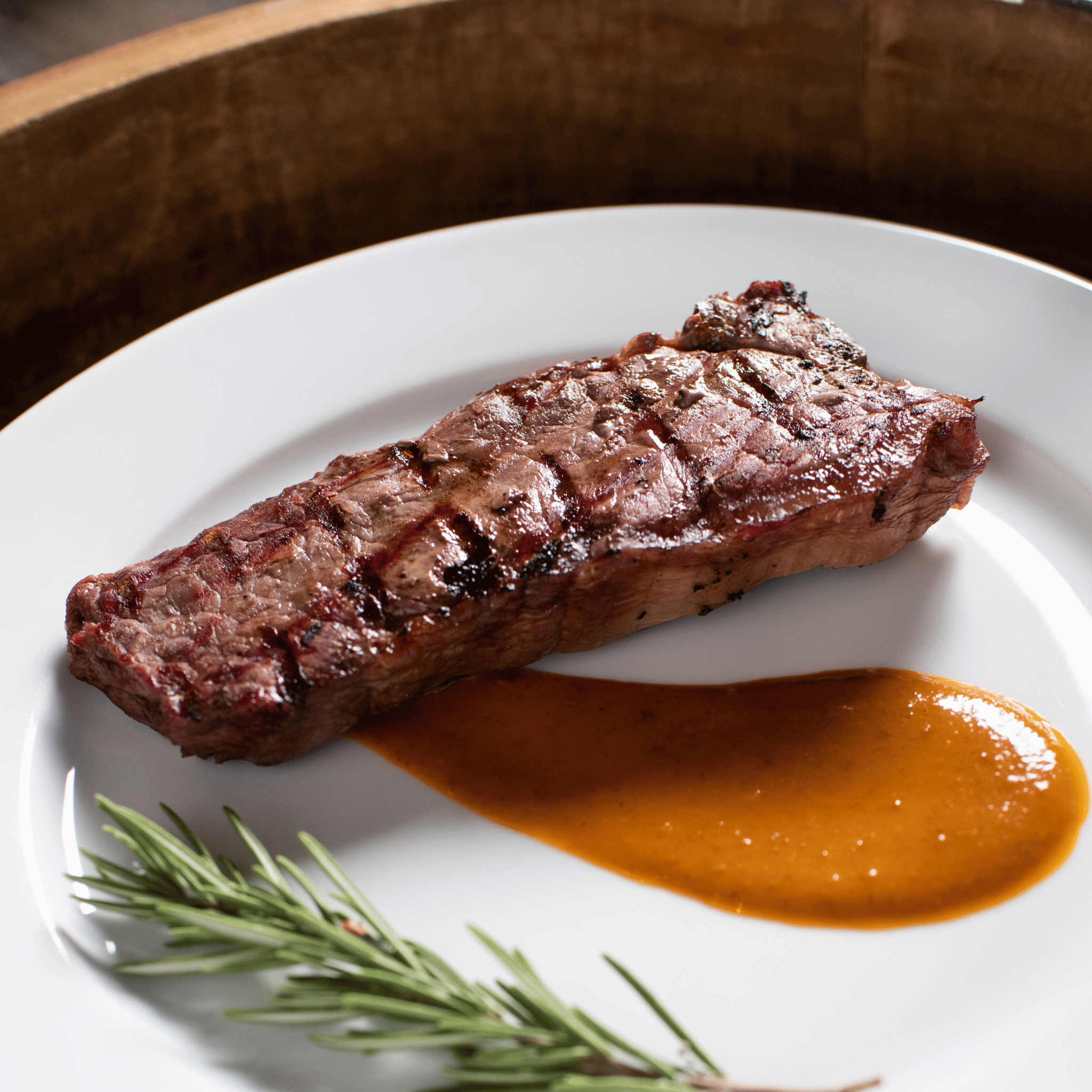 Grilled bison NY strip steak on a white plate with a side of sauce and rosemary.