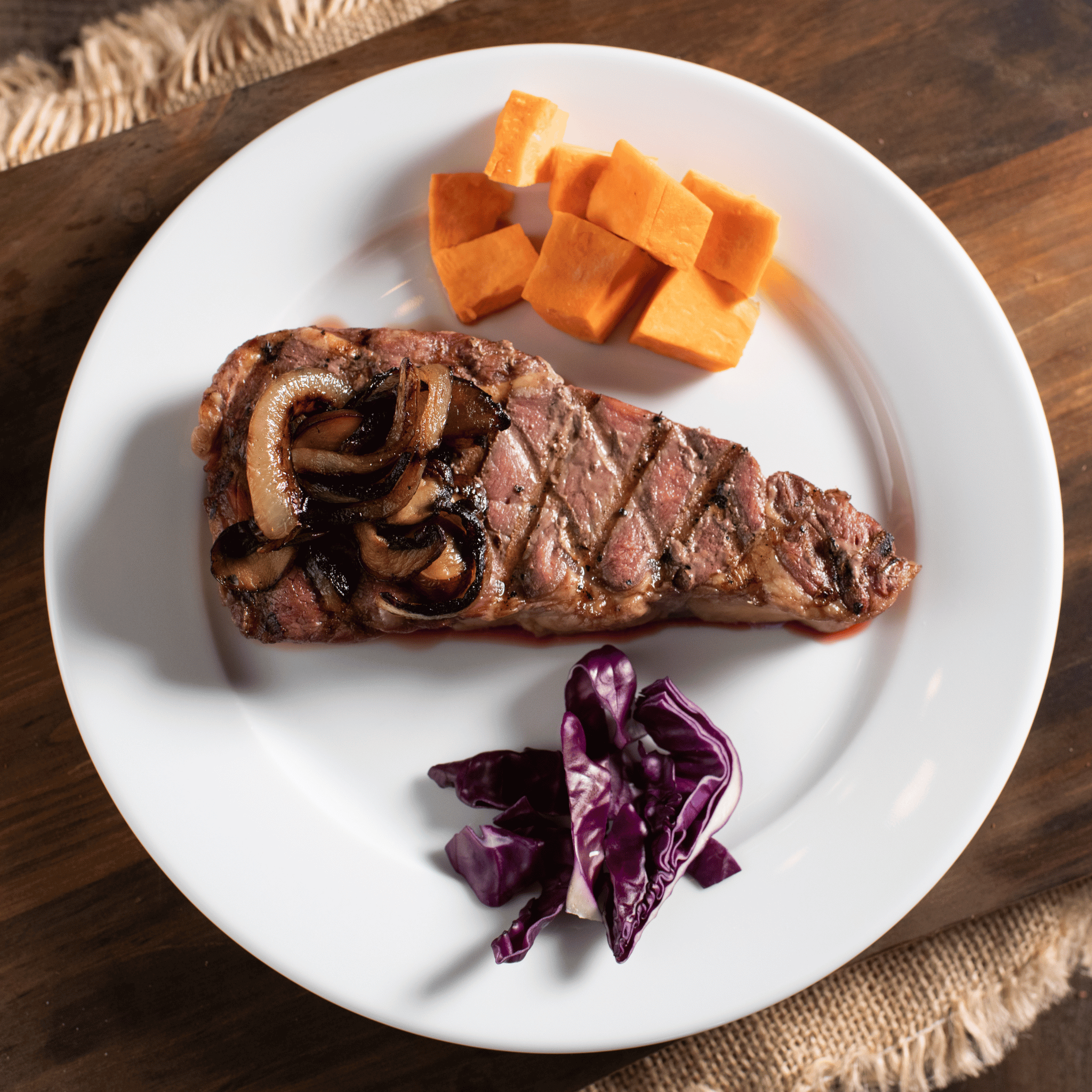 Bison NY Strip Steak with onions, sweet potatoes, and purple cabbage on a white plate.