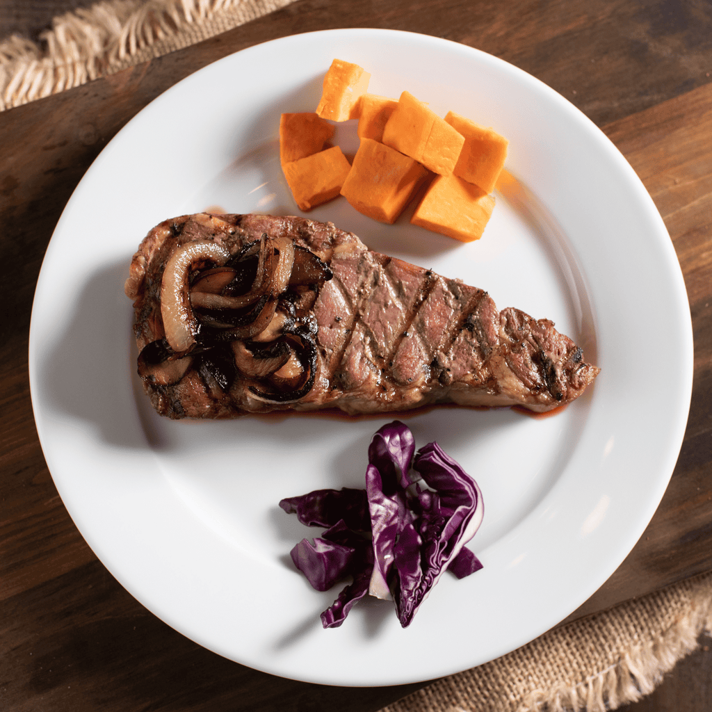 Bison NY Strip Steak with onions, sweet potatoes, and purple cabbage on a white plate.