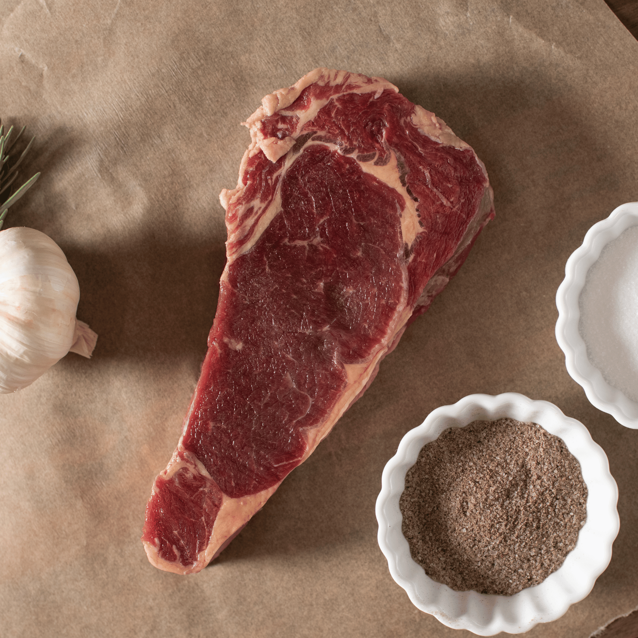 Raw bison NY strip steak on brown paper with a small bowl of seasoning and garlic bulb.