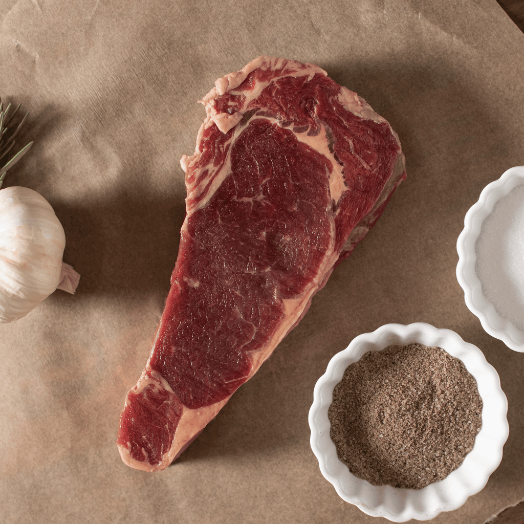 Raw bison NY strip steak on brown paper with a small bowl of seasoning and garlic bulb.