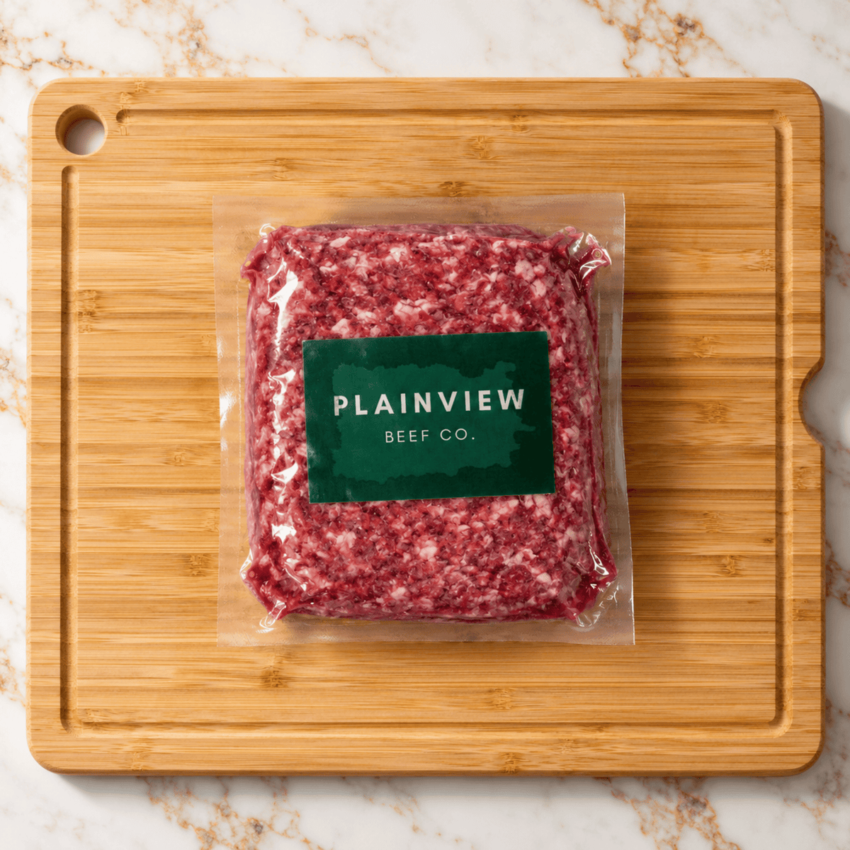 A vacuum-sealed package of ancestral ground beef with a green label reading 'PLAINVIEW BEEF CO.' placed on a rectangular bamboo cutting board over a white marble surface.