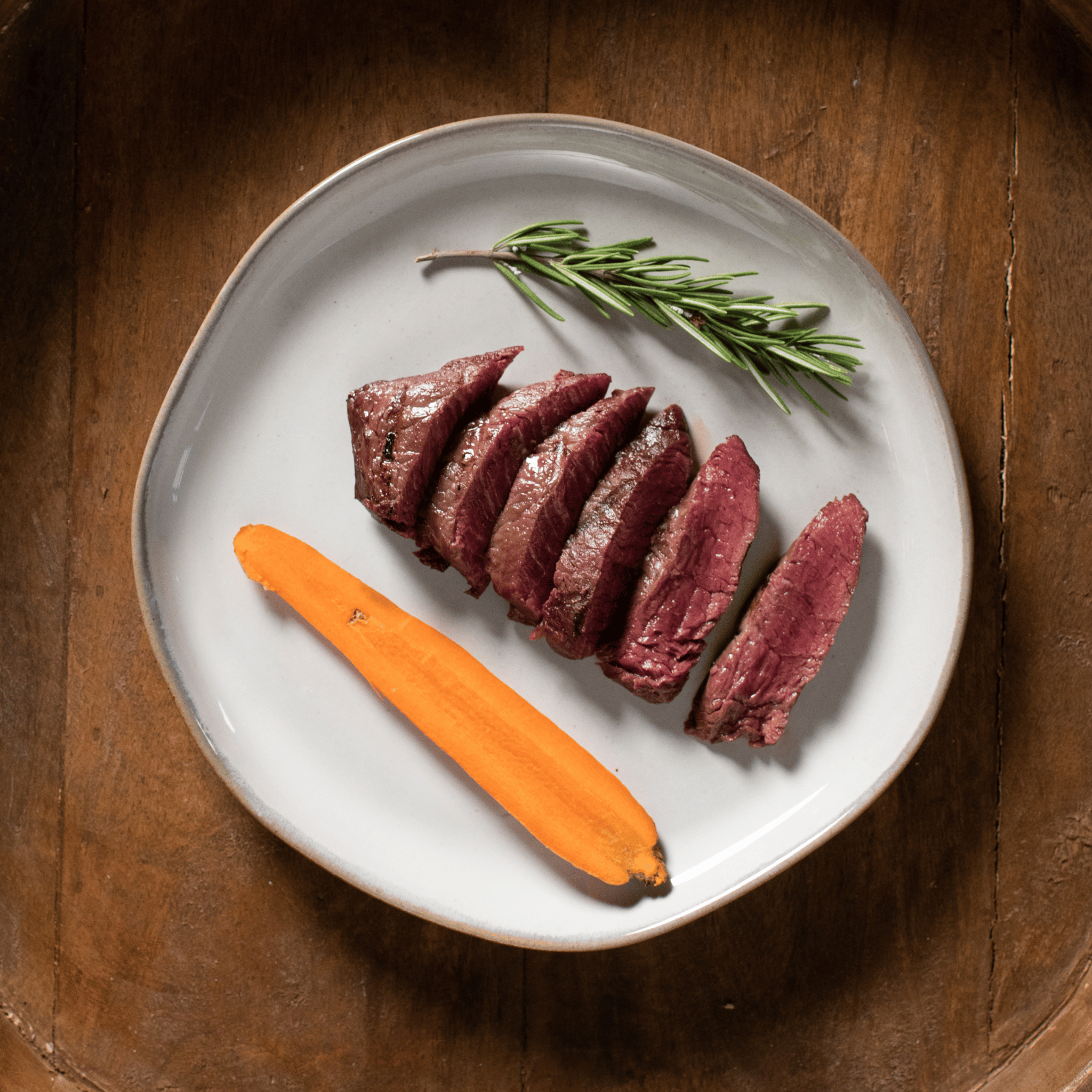 Sliced bison tenderloin filet steak with a carrot and rosemary on a white plate, placed on a wooden surface.