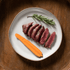 Sliced bison tenderloin filet steak with a carrot and rosemary on a white plate, placed on a wooden surface.