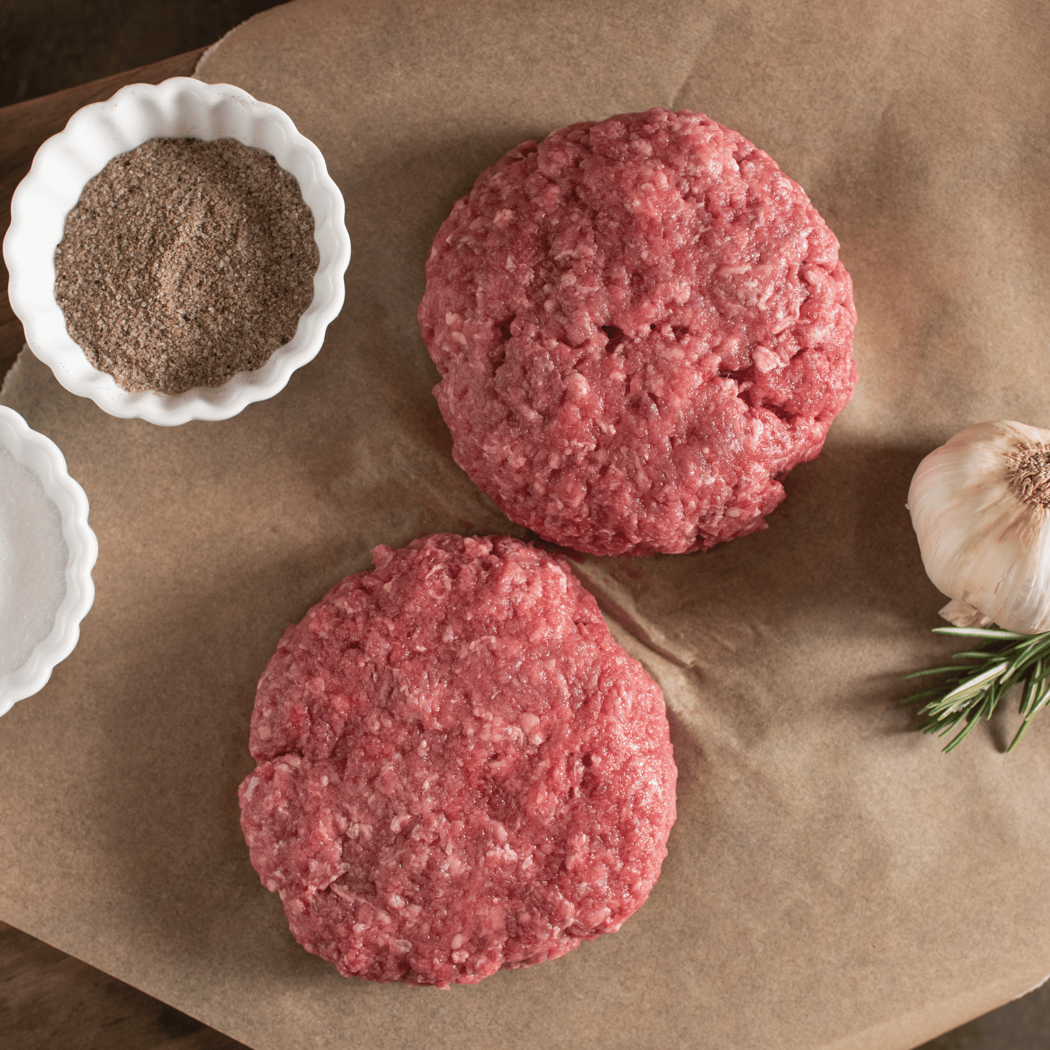 Two raw bison burger patties on brown paper with a small bowl of spices and a clove of garlic.