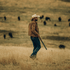 Man in a cowboy hat and leather jacket walking through a field with bison in the background