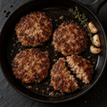 Four browned ground KuneKune pork patties in a black cast-iron skillet with garlic cloves and a sprig of thyme, photographed from above on a dark surface.
