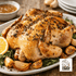 Roasted chicken on a platter with herbs and lemon, served with garlic bulbs and a bowl of sauce.