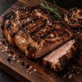 A close-up of a grilled pork shoulder KuneKune steak on a dark wooden cutting board, with one slice cut and placed beside the main piece. Rosemary, a garlic bulb, and coarse salt are visible around the meat.