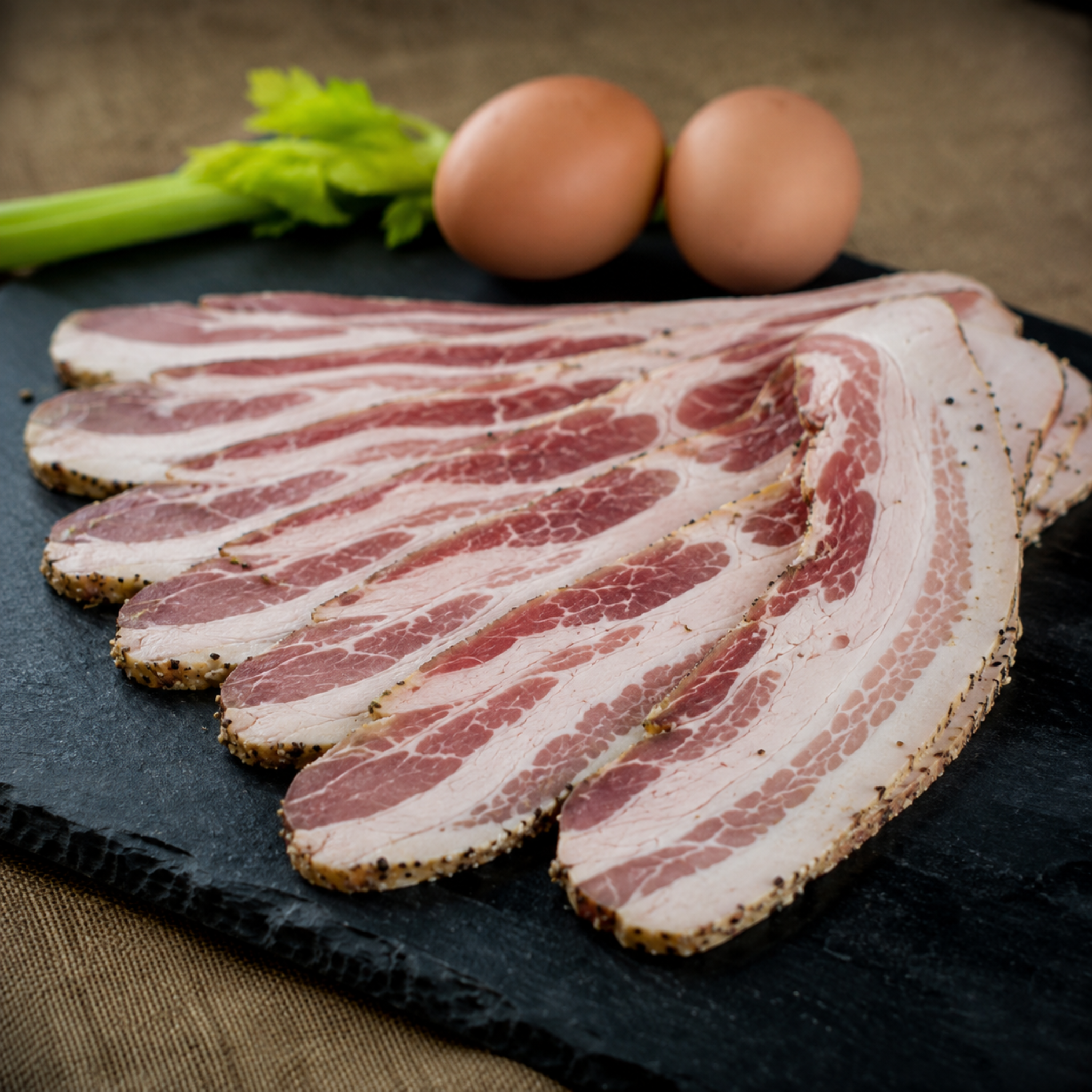 Thin slices of raw bacon arranged on a dark slate board, with two brown eggs and green celery visible in the background on a rustic beige surface.